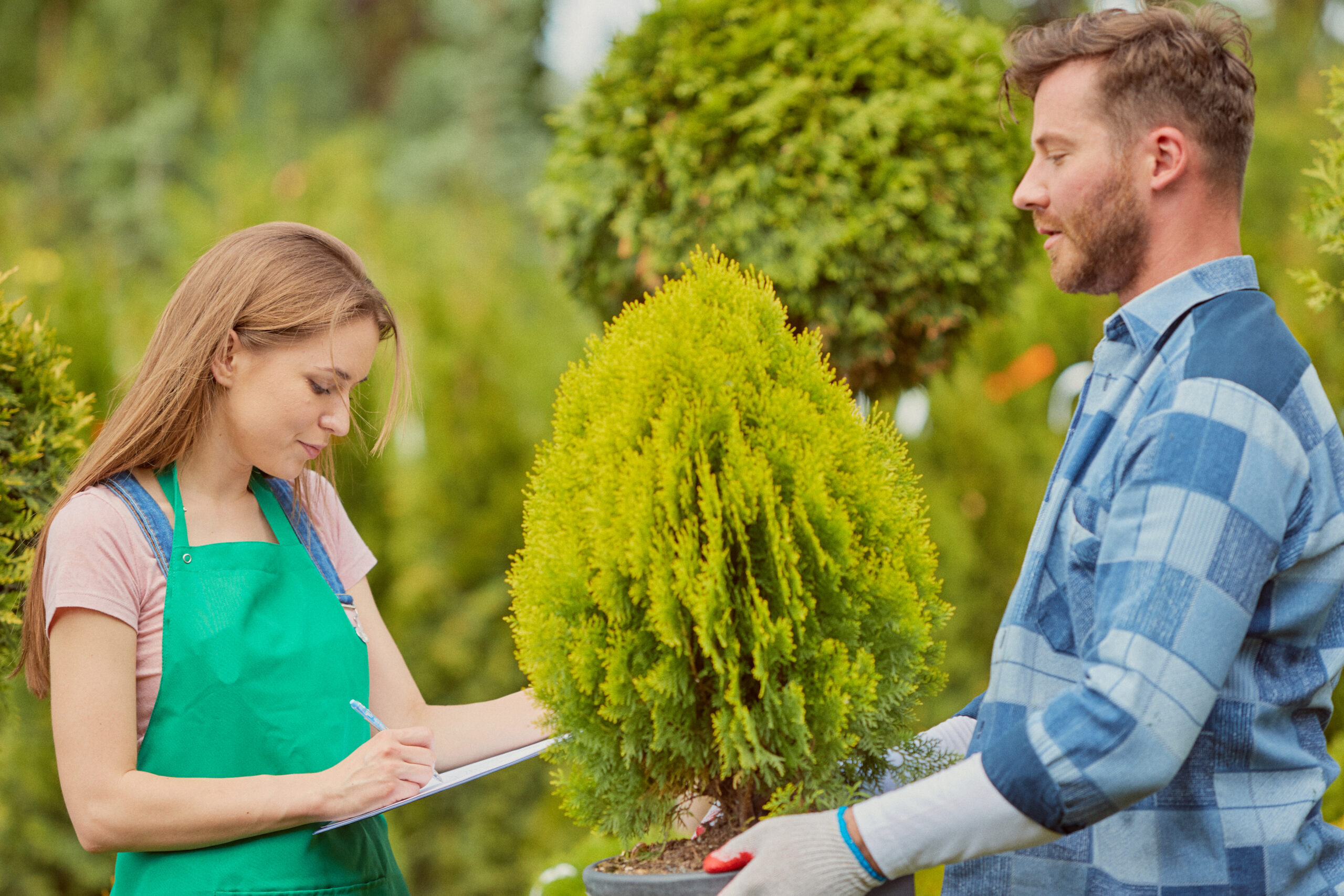 Young gardener woman standing and writing in documents while man doing horticulture job in the garden.